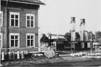 Bilden tagen från Västra vägen.
Till vänster ligger det mellersta och största bostadshuset på glasbrukets område.
Till höger är det äldsta och östligaste bostadshuset under rivning.
Mellan dessa hus skymtar man det s k dårhuset på lasarettets område.