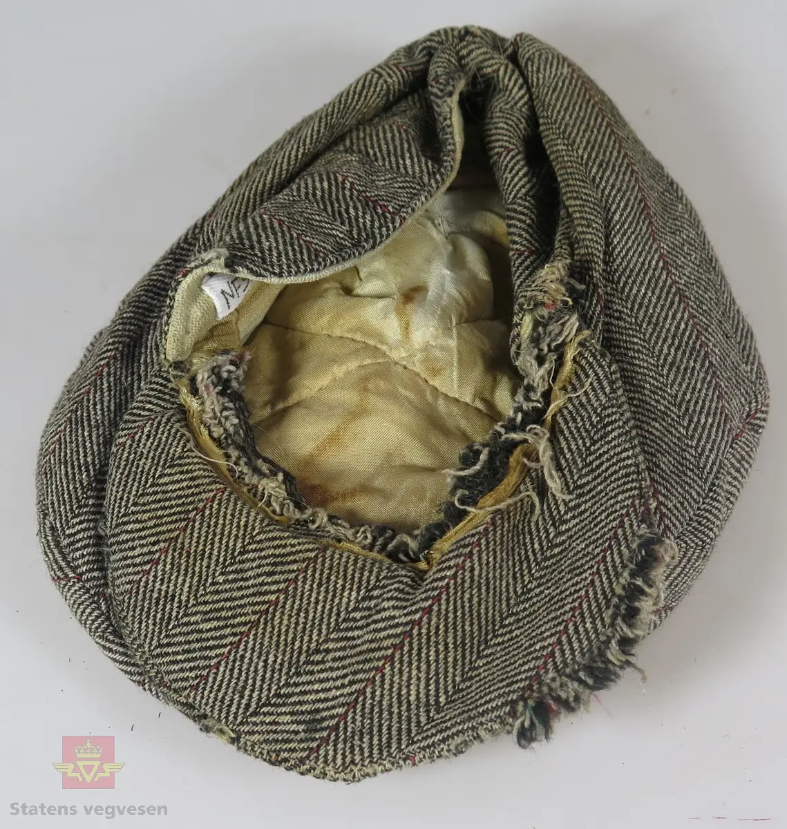 Lua er laget i ullstoff, tweedstoff i fiskebeinsmønster. Hovedfargene er sauebrunt og ubleika med en smal stripe i rødt/rustrød. Skyggen/skjermen er stivet opp med en pappplate. Trykk-knapp holder lua og skyggen sammen. Øreklaffer kan trekkes ned. Vatert silkefôr eggeskallshvit farge. Bl.a. er skyggen noe defekt, dvs med hull. Papplaten i skyggen er delt i to. Lua er vasket etter innkomst til ausjonarius. Lua er ganske slitt.