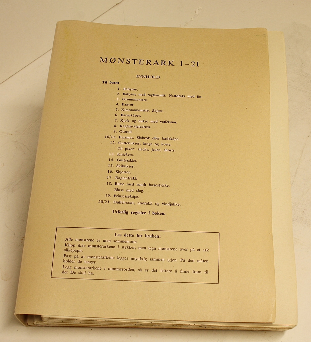 Rektangulær, stående koffert i hvitt og svart med lokk, håndtak på toppen og lås foran. Den inneholder boka "Den Store Syboken" av Ella Lund, samt to mapper med 21 mønsterark i hver med mønster for sying av klær til barn (blå), tenåringer (orange), voksne kvinner (grønn), herrer (svart) og dukker (rød).