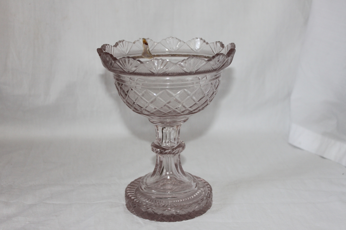 Skål på fot i slipad kristall. Ihålig fot som övergår i ett ben med slipad nod på mitten. Behållaren är rutslipad med solfjäderformad överkant. Lock saknas.