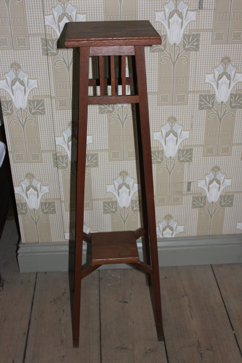 Piedestal i enkel modell, tillverkad i ek. Avsmalnande uppåt. Två fyrkantiga skivor för blomkrukor, en upptill och en nedåt till på benställningen. Den nedre hålls fast av korslagda pinnar. Under den övre skivan finns ett ramverk med lodräta pinnar.