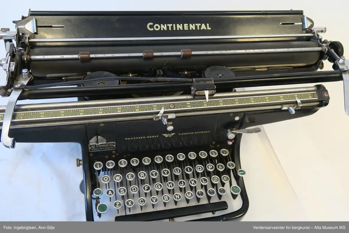 Rektangulær svartlakkert mekanisk skrivemaskin på fire bein med en bred skrivevalse som går langt utover maskinens grunnform. Maskinen har et overtrekk i tekstil.