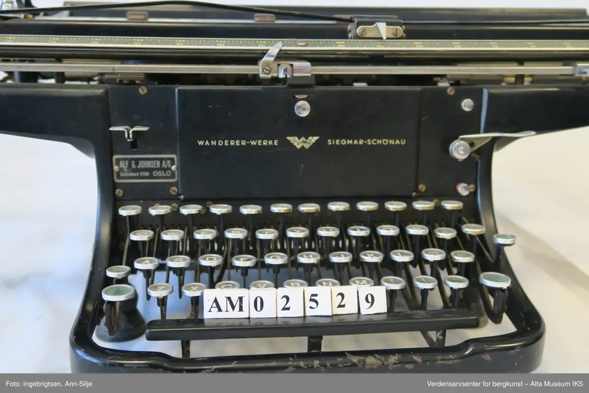 Rektangulær svartlakkert mekanisk skrivemaskin på fire bein med en bred skrivevalse som går langt utover maskinens grunnform. Maskinen har et overtrekk i tekstil.