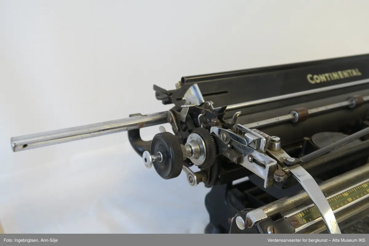 Rektangulær svartlakkert mekanisk skrivemaskin på fire bein med en bred skrivevalse som går langt utover maskinens grunnform. Maskinen har et overtrekk i tekstil.