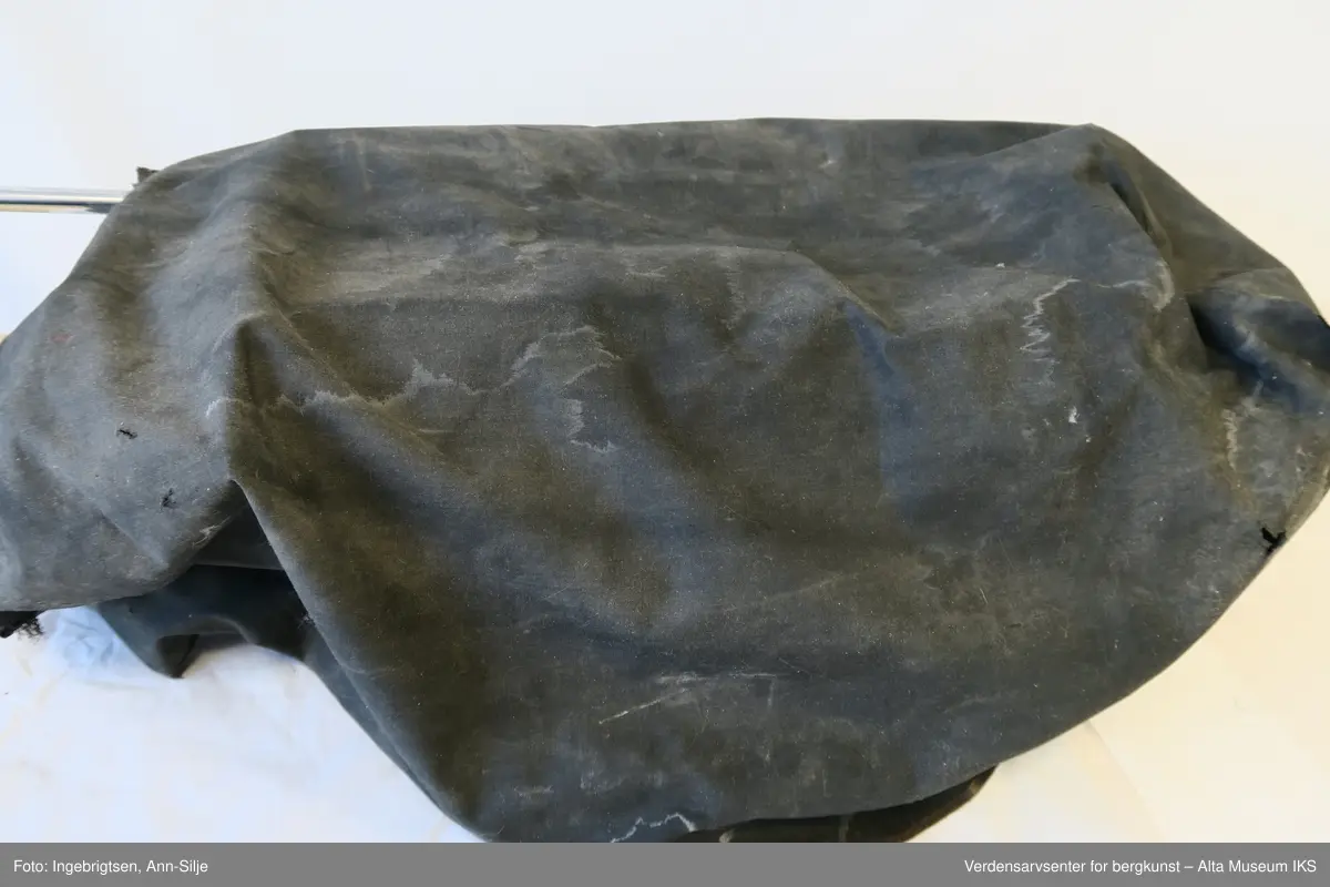 Rektangulær svartlakkert mekanisk skrivemaskin på fire bein med en bred skrivevalse som går langt utover maskinens grunnform. Maskinen har et overtrekk i tekstil.