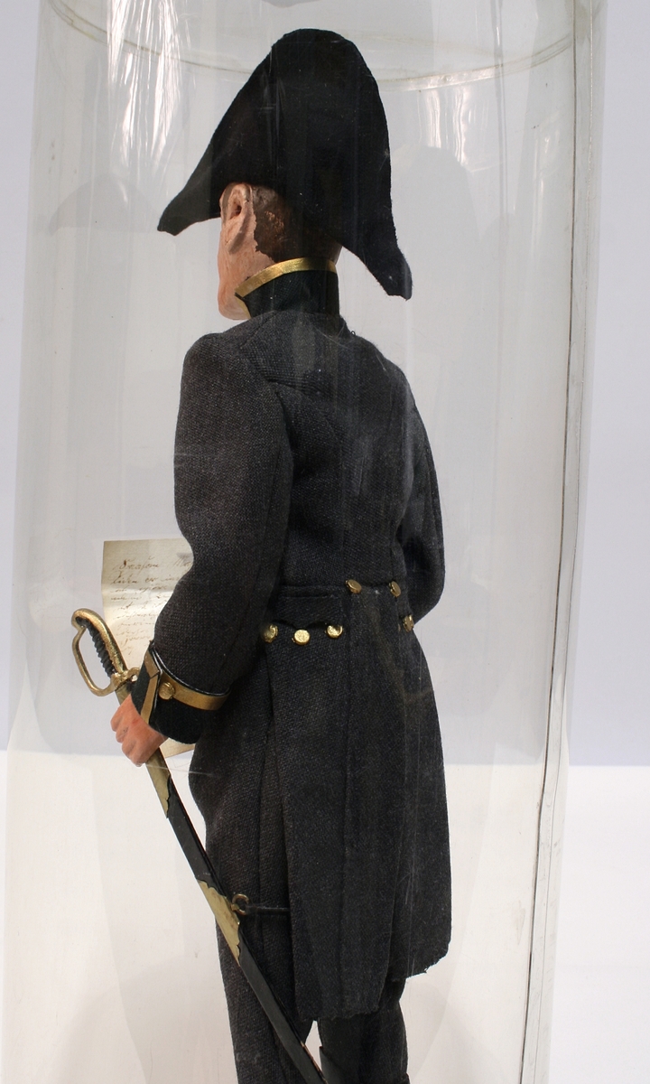 Grå kjole og benklær, grønn krage og oppslag, sorte støvler, sort snutehatt med gullkrampe, sabel med geheng. Montert på sirkelrund plate. Modellen har gradstegn for politiadjutant. Modellen står inni en plastsylinder på en treplate.