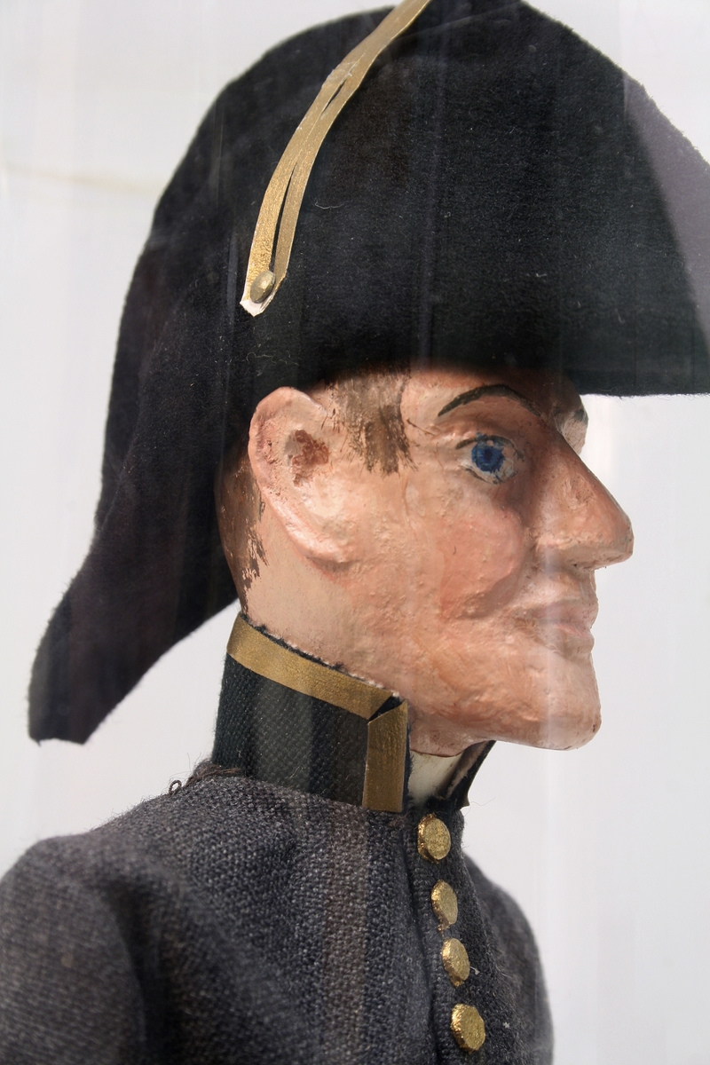 Grå kjole og benklær, grønn krage og oppslag, sorte støvler, sort snutehatt med gullkrampe, sabel med geheng. Montert på sirkelrund plate. Modellen har gradstegn for politiadjutant. Modellen står inni en plastsylinder på en treplate.