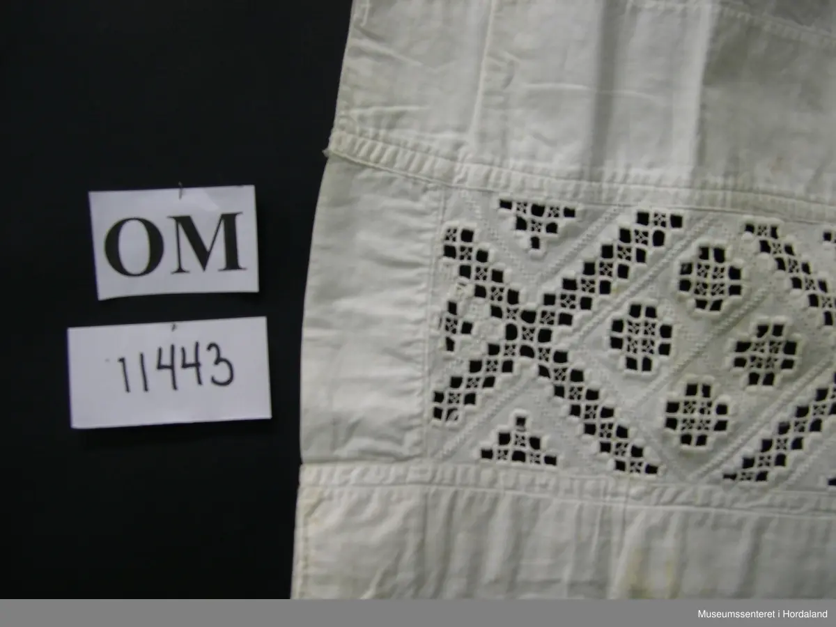 Kvitt bunadsforklede med hardangersaumbord felt inn 14 cm frå nedre kanten. Motivet er inndelt i firkantar, i midten framme er det sydd ei åttebladrose. Linning i livet som fortset rundt ryggen og knappast i sida. Forkledet er rynka ved linninga midt framme. Ikkje rynka i sidene.