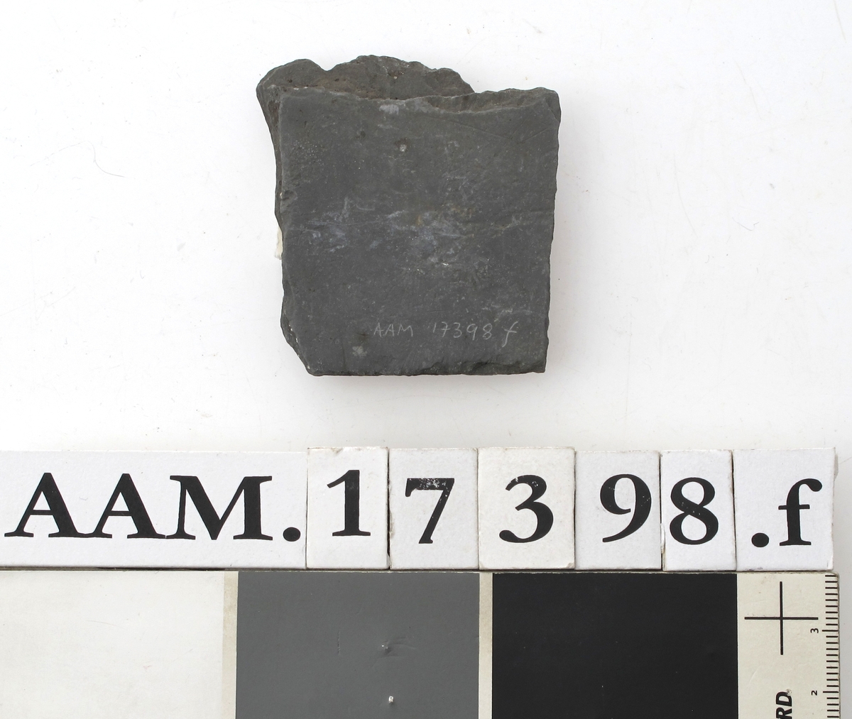 Skifertavle i fire deler .  
a)  17,7 x 8. Femkantet hjørnestykke.  
b)  15 x 6,3. Hjørnestykke med et hakk,  den lengste side er buet. A-b = G 1435.  
c)  16,5 x 5,8. Smalt spisst bladformet. = G 1153.  
d)   Lite skiferstykke. L. 4,8. B. 1,5