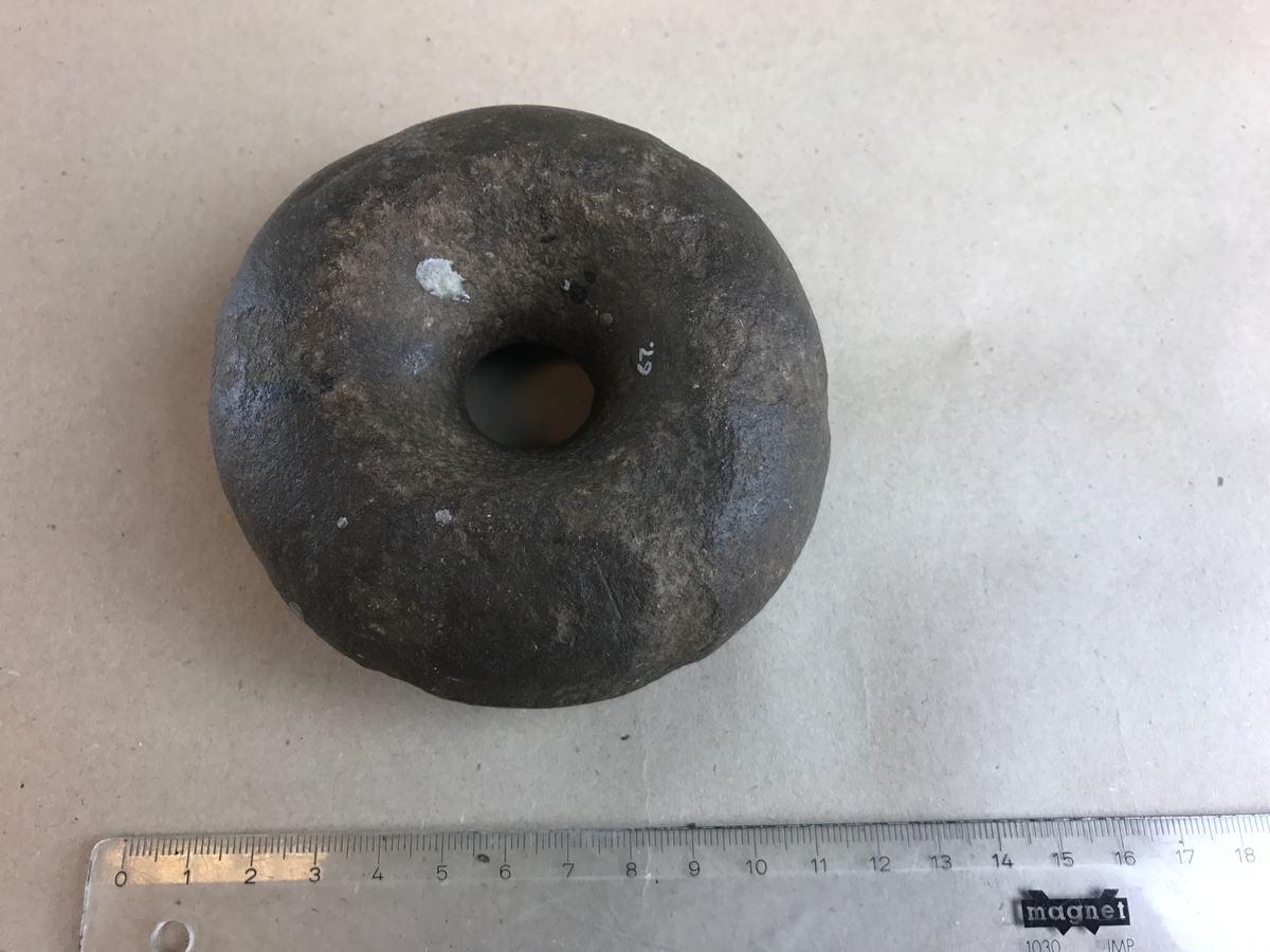 Vävtyngd. Cirkelrund med ett hål, ca 20 mm diam., i mitten. Av kvartsit?