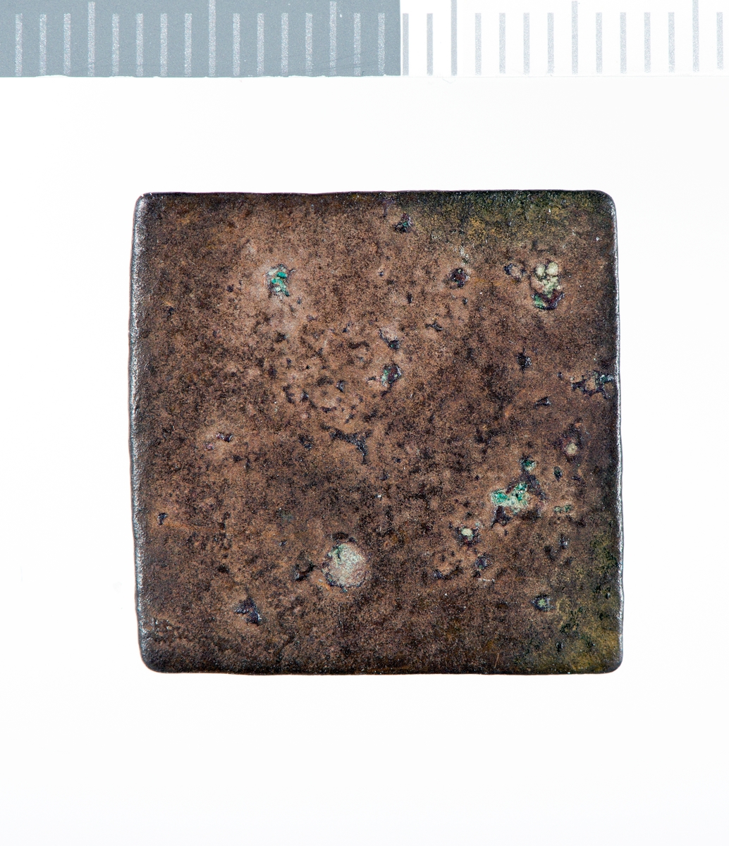 Kvadratisk metallplate med korrodert og  slitt overflate. Sider og hjørner er filt til. Det er rester av et preg på den ene flaten.