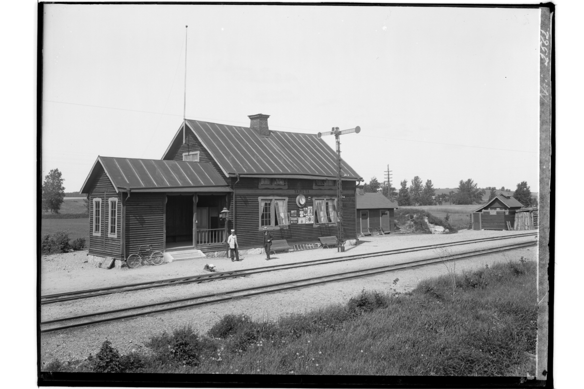 Tarsta Järnvägsstation, envånings stationsbyggnad.
Liten byggnad innehållande toaletter.
2 personer framför byggnaden.