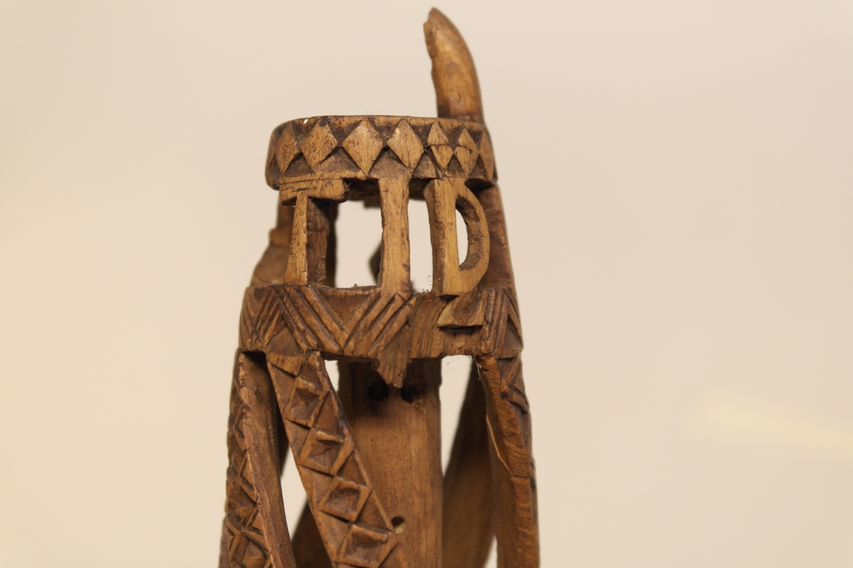 Nøstepinne i skåret furu. Skåret ut med snodde spiler smykket med geometriske mønstre. Øverst på pinnen er det skåret ut, slik at bokstavene står fritt.