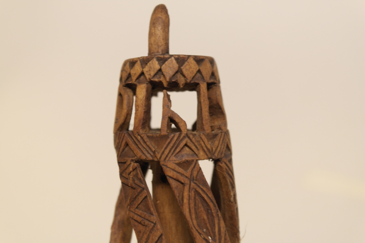 Nøstepinne i skåret furu. Skåret ut med snodde spiler smykket med geometriske mønstre. Øverst på pinnen er det skåret ut, slik at bokstavene står fritt.