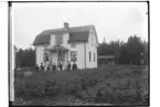 En och en halvplans bostadshus med veranda och frontespis, 10 personer framför huset.
Karl Linden