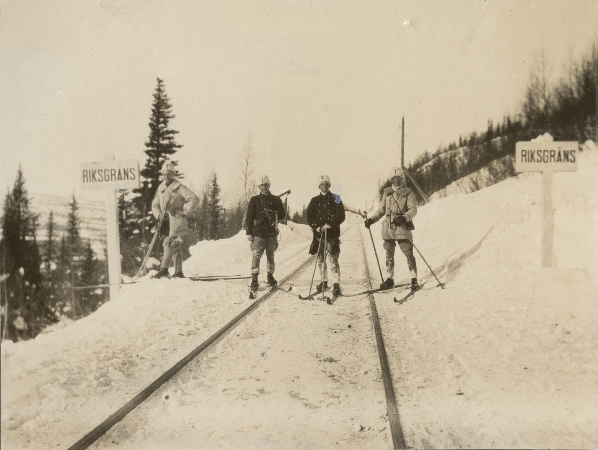 Landsgränsen mellan Sverige och Norge. Fyra soldater på skidor står på järnvägsrälsen mellan två skyltar med text "Riksgräns". Bakom gruppen syns några träd och berg.