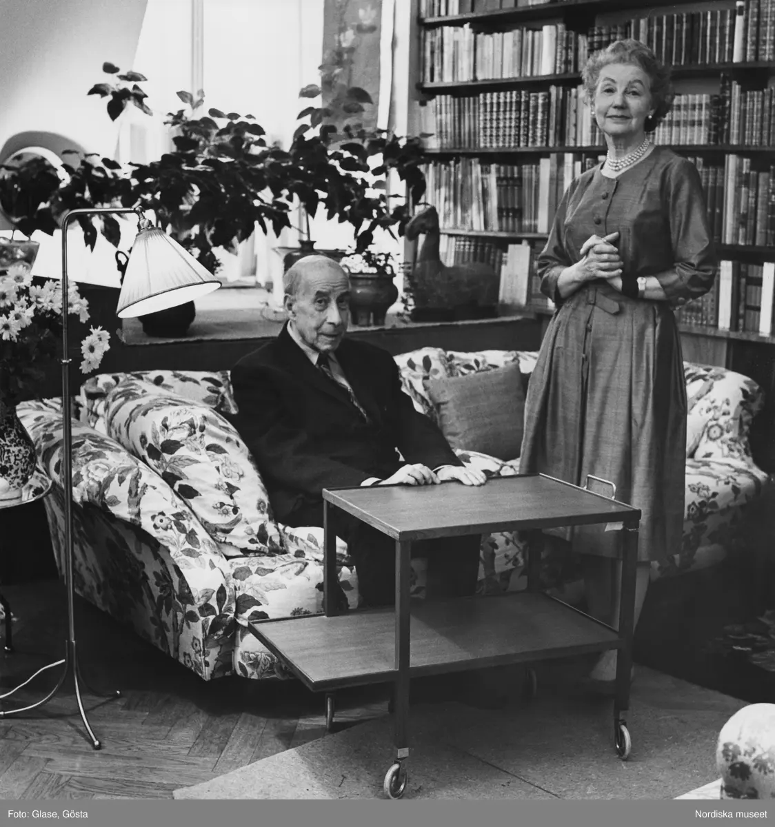 Svenskt Tenns grundare Estrid Ericson och hennes vän och medarbetare arkitekten Josef Frank. Josef Frank sitter i den så kallade Liljevalchssoffan klädd i Bakers tyg Körsbär och visar upp en tevagn han formgivit för Haus & Garten. Troligtvis ett vardagsrum eller sällskapsrum.