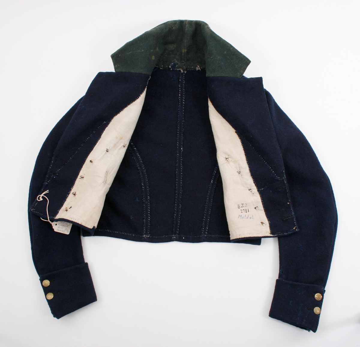 Kort trøye i mørk blått ullstoff, kypertvevd. Overkrage i mørk grønt ullstoff. Underkrage og slagene er mørkeblå. Trøyen er dobbeltspent med buet knappestilling 6 x 2. Den lukkes bare med 2 knapperKnappene er preget med en blomst i midten og en ytterkant av trekanter. På framstykket er det 2lommeklaffer, men bare 1 lomme foret med ubleket linstoff. Kun framstykket er foret. Sidesømmer og skuldersømmer er bakovertrekte. Trange tosømsermer med mansjetter som har 2 messingfargede knapper og 2 knapphull. Stikninger rundt kragen, slagene, MF og nedre kant av trøyen. Høyre undererme er skjøtt oppe ved ermhulen og nede ved mansjetten.