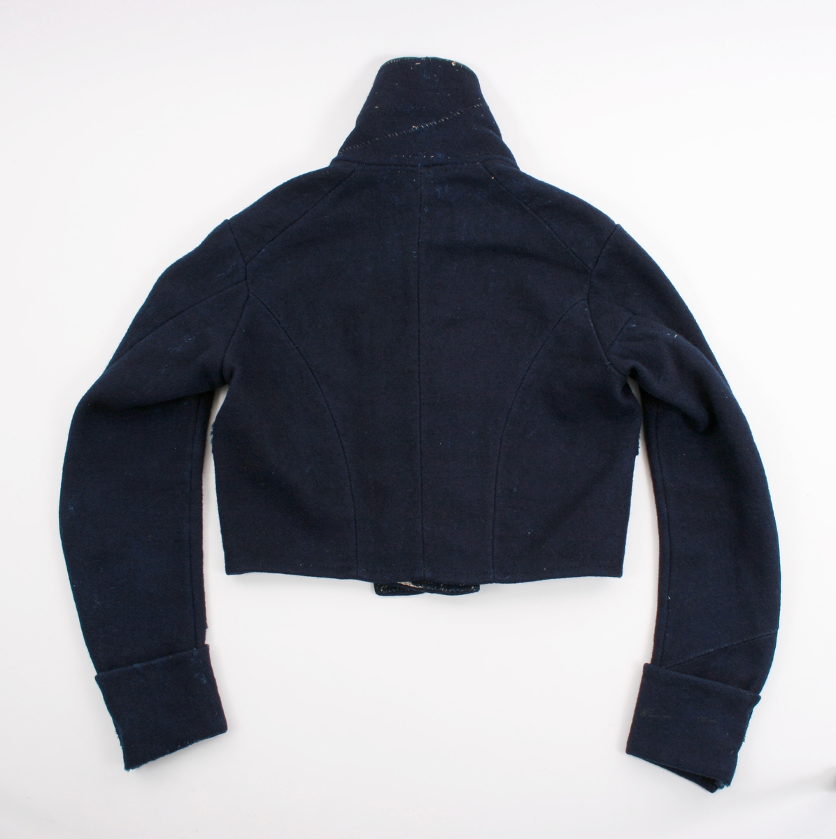 Kort trøye i mørk blått ullstoff, kypertvevd. Overkrage i mørk grønt ullstoff. Underkrage og slagene er mørkeblå. Trøyen er dobbeltspent med buet knappestilling 6 x 2. Den lukkes bare med 2 knapperKnappene er preget med en blomst i midten og en ytterkant av trekanter. På framstykket er det 2lommeklaffer, men bare 1 lomme foret med ubleket linstoff. Kun framstykket er foret. Sidesømmer og skuldersømmer er bakovertrekte. Trange tosømsermer med mansjetter som har 2 messingfargede knapper og 2 knapphull. Stikninger rundt kragen, slagene, MF og nedre kant av trøyen. Høyre undererme er skjøtt oppe ved ermhulen og nede ved mansjetten.