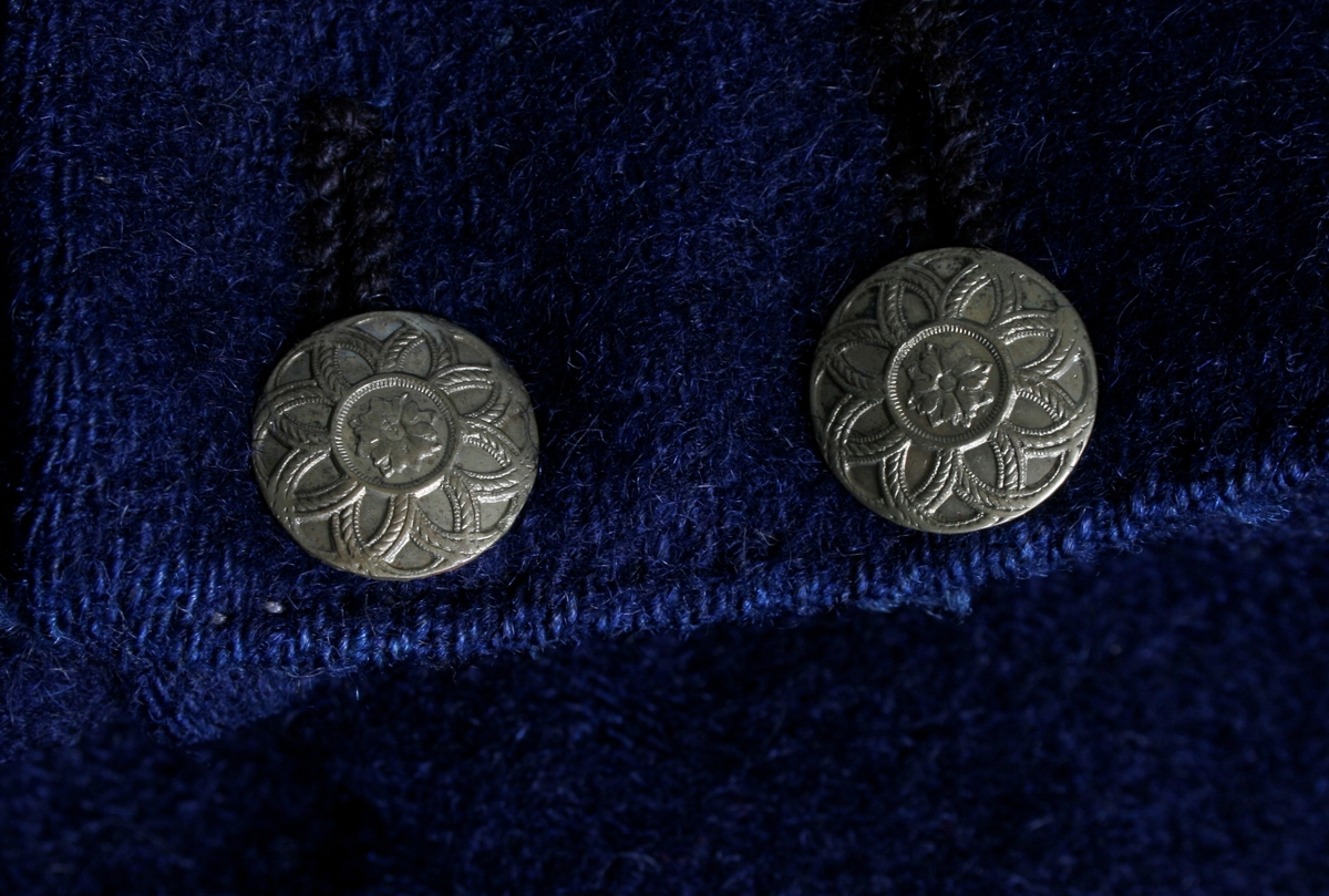 Kort trøye i mørk blått ullstoff, kypertvevd. Overkrage i mørk grønt ullstoff. Underkrage og slagene er mørkeblå. Trøyen er dobbeltspent med buet knappestilling 6 x 2. Den lukkes bare med 2 knapperKnappene er preget med en blomst i midten og en ytterkant av trekanter. På framstykket er det 2lommeklaffer, men bare 1 lomme foret med ubleket linstoff. Kun framstykket er foret. Sidesømmer og skuldersømmer er bakovertrekte. Trange tosømsermer med mansjetter som har 2 messingfargede knapper og 2 knapphull. Stikninger rundt kragen, slagene, MF og nedre kant av trøyen. Høyre undererme er skjøtt oppe ved ermhulen og nede ved mansjetten.