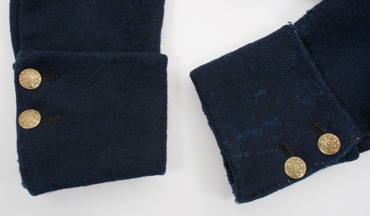 Kort trøye i mørk blått ullstoff, kypertvevd. Overkrage i mørk grønt ullstoff. Underkrage og slagene er mørkeblå. Trøyen er dobbeltspent med buet knappestilling 6 x 2. Den lukkes bare med 2 knapperKnappene er preget med en blomst i midten og en ytterkant av trekanter. På framstykket er det 2lommeklaffer, men bare 1 lomme foret med ubleket linstoff. Kun framstykket er foret. Sidesømmer og skuldersømmer er bakovertrekte. Trange tosømsermer med mansjetter som har 2 messingfargede knapper og 2 knapphull. Stikninger rundt kragen, slagene, MF og nedre kant av trøyen. Høyre undererme er skjøtt oppe ved ermhulen og nede ved mansjetten.