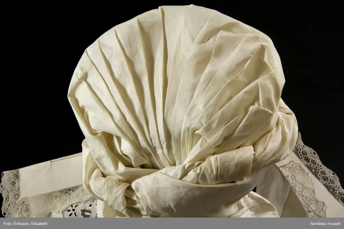 b. Brudtröja av svart vadmal,  Runt alla kanter bård av ytterst brunsvart sammetsband och innanför ett 4 cm brett svart sammetsband  med invävt bladmönster. På ärmarna ett svart slätt sammetsband med pålagd guldspets därefter siden band i rosa och grönt på botten av  svart  sammet och överst i bården en knypplad guldspets. 
c: Förkläde. Material: Textil, bomull. Teknik: Manufakturvävning. Handsömnad. Längd: 77 cm. Bredd: 110 cm. Färg: Vit.
Förkläde i vitt manufakturvävt bomullstyg, ihopsytt i mitten, smalrandigt, stärkt. Rynkningar mot midjelinning i tuskaftat bomullstyg, 1,5 cm bred, fastsydda bomullsband i sidorna. Uppvikt fåll nedtill, 6,5 cm, ovanför fåll, 5 lagda veck med ca 1 cm mellanrum, enkel utdragssöm i kanten av vecken.
/Inga-Lill Eliasson 2008-02-25

e: Krage. Längd: 47,5, cm - totallängd, bröststycke. Bredd: 33 cm - bröststycke. Höjd: 14 cm - kraghöjd, inkl spets. Material: Textil, bomull. Lingarn - spets. Bomullsgarn - broderi. Teknik: Tuskaft. Vitbroderi, Knyppling. Handsömnad.
Krage i tuskaftat vitt bomullstyg. Rektangulär duk, uppklippt halshål med fastsydd krage, 14 cm hög. Kragen har 3,5 cm bred fåll, enkel utdragssöm i kanten. Vitbroderi  i framkant,  plattsöm och kantsydda hål med infällt tyll, två sydda knapphål. Handknypplad spets fastsydd på långsida och korstsidor, 2,5 cm bred. Öppning fram, 24 cm långt sprund. Kragen är handsydd.
Lästips: Svensson Sigfrid, (1935). Skånes Folkdräkter, en dräkthistorisk undersökning 1500 - 1900. Stockholm: Nordiska Museets Handlingar: 3.
/Inga-Lill Eliasson 2008-02-19