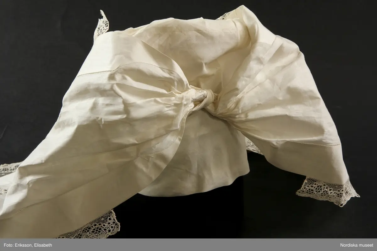 b. Brudtröja av svart vadmal,  Runt alla kanter bård av ytterst brunsvart sammetsband och innanför ett 4 cm brett svart sammetsband  med invävt bladmönster. På ärmarna ett svart slätt sammetsband med pålagd guldspets därefter siden band i rosa och grönt på botten av  svart  sammet och överst i bården en knypplad guldspets. 
c: Förkläde. Material: Textil, bomull. Teknik: Manufakturvävning. Handsömnad. Längd: 77 cm. Bredd: 110 cm. Färg: Vit.
Förkläde i vitt manufakturvävt bomullstyg, ihopsytt i mitten, smalrandigt, stärkt. Rynkningar mot midjelinning i tuskaftat bomullstyg, 1,5 cm bred, fastsydda bomullsband i sidorna. Uppvikt fåll nedtill, 6,5 cm, ovanför fåll, 5 lagda veck med ca 1 cm mellanrum, enkel utdragssöm i kanten av vecken.
/Inga-Lill Eliasson 2008-02-25

e: Krage. Längd: 47,5, cm - totallängd, bröststycke. Bredd: 33 cm - bröststycke. Höjd: 14 cm - kraghöjd, inkl spets. Material: Textil, bomull. Lingarn - spets. Bomullsgarn - broderi. Teknik: Tuskaft. Vitbroderi, Knyppling. Handsömnad.
Krage i tuskaftat vitt bomullstyg. Rektangulär duk, uppklippt halshål med fastsydd krage, 14 cm hög. Kragen har 3,5 cm bred fåll, enkel utdragssöm i kanten. Vitbroderi  i framkant,  plattsöm och kantsydda hål med infällt tyll, två sydda knapphål. Handknypplad spets fastsydd på långsida och korstsidor, 2,5 cm bred. Öppning fram, 24 cm långt sprund. Kragen är handsydd.
Lästips: Svensson Sigfrid, (1935). Skånes Folkdräkter, en dräkthistorisk undersökning 1500 - 1900. Stockholm: Nordiska Museets Handlingar: 3.
/Inga-Lill Eliasson 2008-02-19