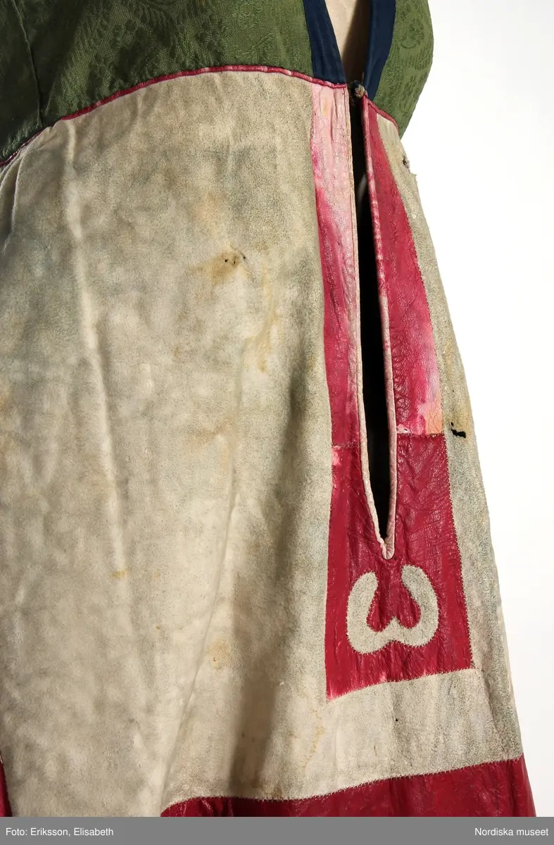 Livkjol, s.k. päls eller skinnklocka. Röd kjol,  där övre halvan består av rött saffianskinn med 7 centimeter bred vit kant överst, och ett vitt fält mitt fram,  nedre halvan av röd vadmal, 3 våder  hela kjolen fodrad med fårskinnspäls och nedtill oblekt linnelärft och är mycket tung. Täta stripade rynkor runtom i midjan utom ett slätt parti 15 cm på var sida om mittsprundet fram. Nedanför sprundet mönster i applikation med vittt skinn på rött . Livstycket är av mönstervävt grönt kamgarnsylle med flotterande blommönster på tuskaftsbotten. 2 framstycken, 1 ryggstycke, kantat med indigo blå 2,5 cm breda bomullsband kring ärmhål hals, framkanter, i vinklar på ryggen blå sidenband.  Livet fodrat med  linnelärft.  En hake och hyska i mässing i midjan, för övrigt ingen knäppning. Har sannolikt snörts fram genom maljor, troligen med en silverkedja.
Det gröna mönstrade tyget var vanligt runt 1800, troligen en importvara från England. Fanns i olika färger.
Se Österlens Folkdräkter, 1999:  Christina Lindwall-Nordin:  Bredhöftade matronor i tjocka kjolar, sid 56-59.
/Berit Eldvik 2012-08-14