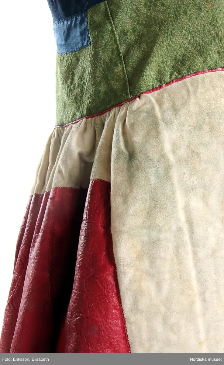 Livkjol, s.k. päls eller skinnklocka. Röd kjol,  där övre halvan består av rött saffianskinn med 7 centimeter bred vit kant överst, och ett vitt fält mitt fram,  nedre halvan av röd vadmal, 3 våder  hela kjolen fodrad med fårskinnspäls och nedtill oblekt linnelärft och är mycket tung. Täta stripade rynkor runtom i midjan utom ett slätt parti 15 cm på var sida om mittsprundet fram. Nedanför sprundet mönster i applikation med vittt skinn på rött . Livstycket är av mönstervävt grönt kamgarnsylle med flotterande blommönster på tuskaftsbotten. 2 framstycken, 1 ryggstycke, kantat med indigo blå 2,5 cm breda bomullsband kring ärmhål hals, framkanter, i vinklar på ryggen blå sidenband.  Livet fodrat med  linnelärft.  En hake och hyska i mässing i midjan, för övrigt ingen knäppning. Har sannolikt snörts fram genom maljor, troligen med en silverkedja.
Det gröna mönstrade tyget var vanligt runt 1800, troligen en importvara från England. Fanns i olika färger.
Se Österlens Folkdräkter, 1999:  Christina Lindwall-Nordin:  Bredhöftade matronor i tjocka kjolar, sid 56-59.
/Berit Eldvik 2012-08-14