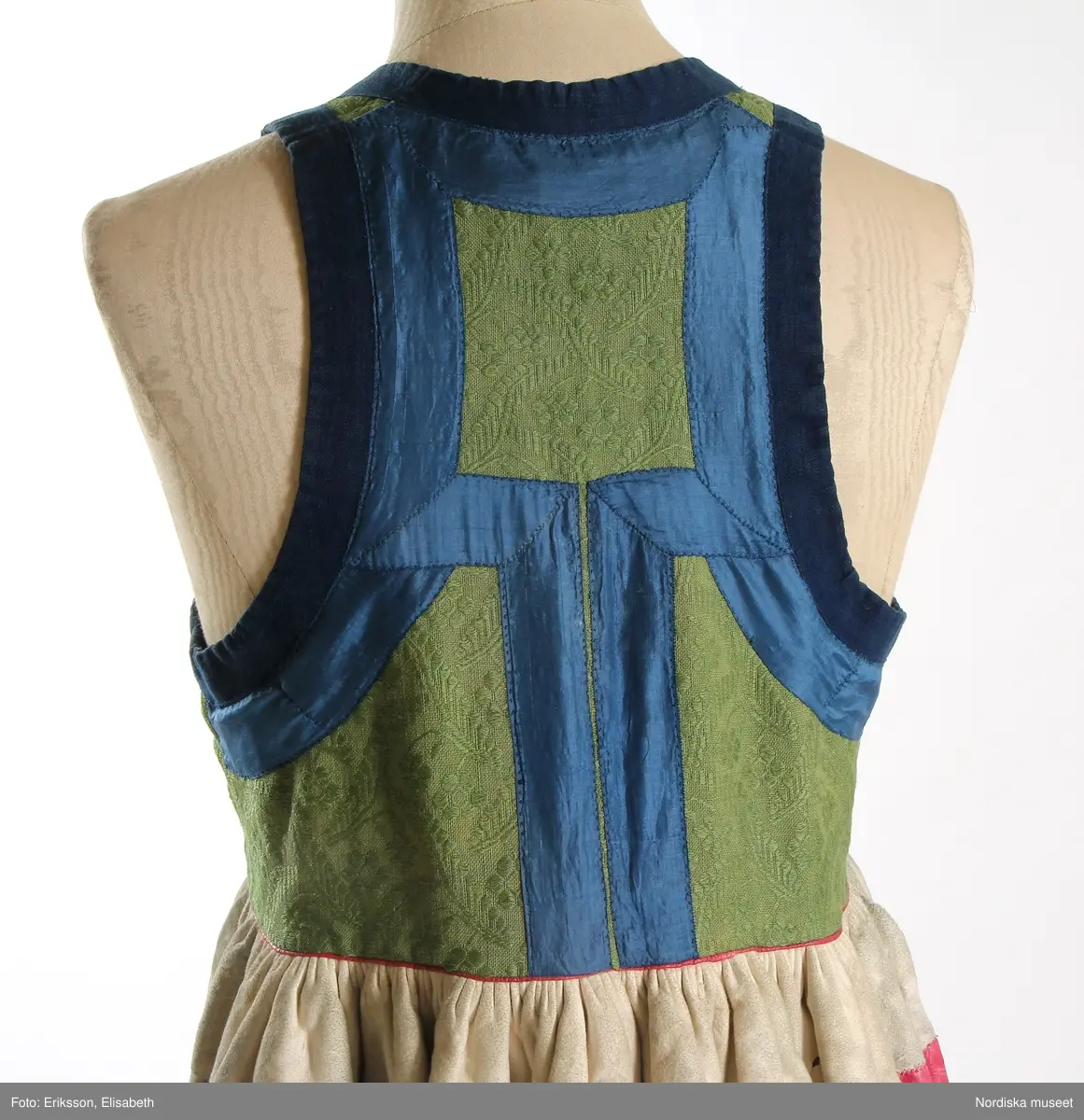 Livkjol, s.k. päls eller skinnklocka. Röd kjol,  där övre halvan består av rött saffianskinn med 7 centimeter bred vit kant överst, och ett vitt fält mitt fram,  nedre halvan av röd vadmal, 3 våder  hela kjolen fodrad med fårskinnspäls och nedtill oblekt linnelärft och är mycket tung. Täta stripade rynkor runtom i midjan utom ett slätt parti 15 cm på var sida om mittsprundet fram. Nedanför sprundet mönster i applikation med vittt skinn på rött . Livstycket är av mönstervävt grönt kamgarnsylle med flotterande blommönster på tuskaftsbotten. 2 framstycken, 1 ryggstycke, kantat med indigo blå 2,5 cm breda bomullsband kring ärmhål hals, framkanter, i vinklar på ryggen blå sidenband.  Livet fodrat med  linnelärft.  En hake och hyska i mässing i midjan, för övrigt ingen knäppning. Har sannolikt snörts fram genom maljor, troligen med en silverkedja.
Det gröna mönstrade tyget var vanligt runt 1800, troligen en importvara från England. Fanns i olika färger.
Se Österlens Folkdräkter, 1999:  Christina Lindwall-Nordin:  Bredhöftade matronor i tjocka kjolar, sid 56-59.
/Berit Eldvik 2012-08-14