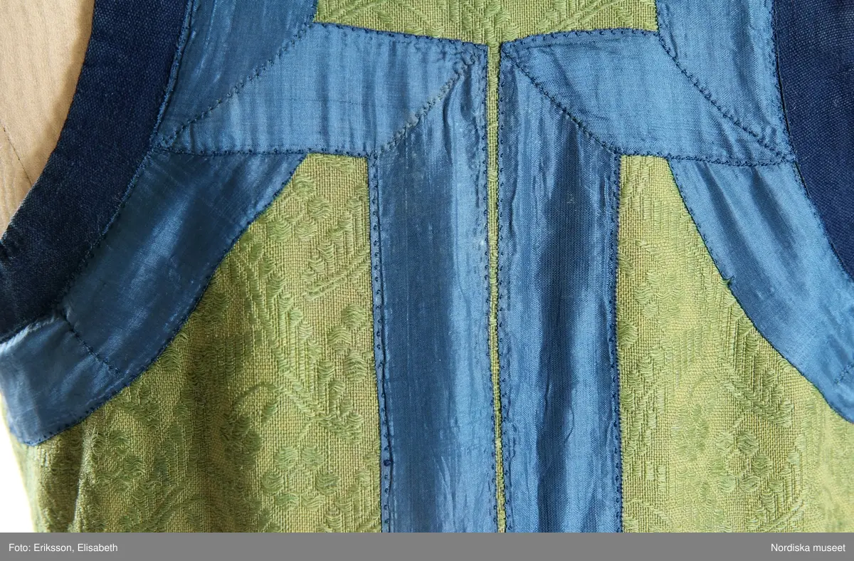 Livkjol, s.k. päls eller skinnklocka. Röd kjol,  där övre halvan består av rött saffianskinn med 7 centimeter bred vit kant överst, och ett vitt fält mitt fram,  nedre halvan av röd vadmal, 3 våder  hela kjolen fodrad med fårskinnspäls och nedtill oblekt linnelärft och är mycket tung. Täta stripade rynkor runtom i midjan utom ett slätt parti 15 cm på var sida om mittsprundet fram. Nedanför sprundet mönster i applikation med vittt skinn på rött . Livstycket är av mönstervävt grönt kamgarnsylle med flotterande blommönster på tuskaftsbotten. 2 framstycken, 1 ryggstycke, kantat med indigo blå 2,5 cm breda bomullsband kring ärmhål hals, framkanter, i vinklar på ryggen blå sidenband.  Livet fodrat med  linnelärft.  En hake och hyska i mässing i midjan, för övrigt ingen knäppning. Har sannolikt snörts fram genom maljor, troligen med en silverkedja.
Det gröna mönstrade tyget var vanligt runt 1800, troligen en importvara från England. Fanns i olika färger.
Se Österlens Folkdräkter, 1999:  Christina Lindwall-Nordin:  Bredhöftade matronor i tjocka kjolar, sid 56-59.
/Berit Eldvik 2012-08-14