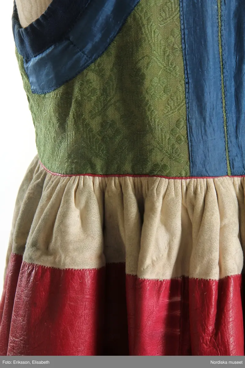 Livkjol, s.k. päls eller skinnklocka. Röd kjol,  där övre halvan består av rött saffianskinn med 7 centimeter bred vit kant överst, och ett vitt fält mitt fram,  nedre halvan av röd vadmal, 3 våder  hela kjolen fodrad med fårskinnspäls och nedtill oblekt linnelärft och är mycket tung. Täta stripade rynkor runtom i midjan utom ett slätt parti 15 cm på var sida om mittsprundet fram. Nedanför sprundet mönster i applikation med vittt skinn på rött . Livstycket är av mönstervävt grönt kamgarnsylle med flotterande blommönster på tuskaftsbotten. 2 framstycken, 1 ryggstycke, kantat med indigo blå 2,5 cm breda bomullsband kring ärmhål hals, framkanter, i vinklar på ryggen blå sidenband.  Livet fodrat med  linnelärft.  En hake och hyska i mässing i midjan, för övrigt ingen knäppning. Har sannolikt snörts fram genom maljor, troligen med en silverkedja.
Det gröna mönstrade tyget var vanligt runt 1800, troligen en importvara från England. Fanns i olika färger.
Se Österlens Folkdräkter, 1999:  Christina Lindwall-Nordin:  Bredhöftade matronor i tjocka kjolar, sid 56-59.
/Berit Eldvik 2012-08-14
