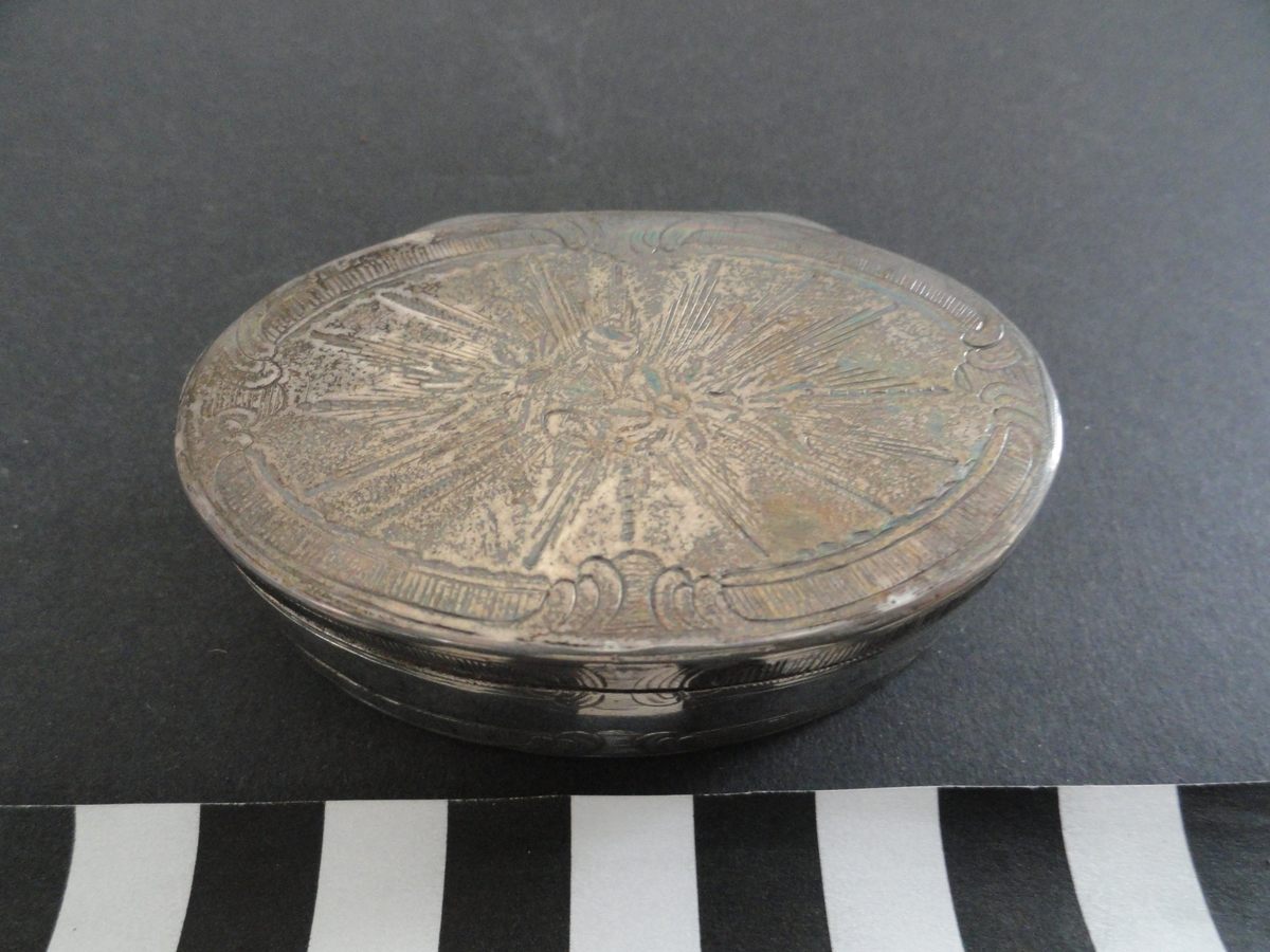 Oval snusdosa av silver. Invändigt förgylld.
Lock med gångjärn.
Graverad dekor i form av blombuketter och stjärnformig strålkrans på lock och botten. Bård av räfflade voluter på lock, sidor och botten.