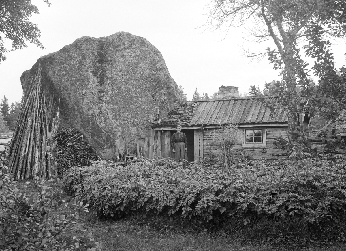 Karolina Jonsdotter utanför sitt undantag på torpet Degerhult i Ydre. Karolina hade tidigare varit torpare på Degerhult tillsammans med sin make Ludwig Karlsson. Efter att denne gått bort 1891 fick hon flytta ut till den avbildade stugan, byggd intill ett flyttblock. Här bodde hon till 1912, de första åren tillsammans med sonen Karl August. Hon avled på fattighuset 1915.
