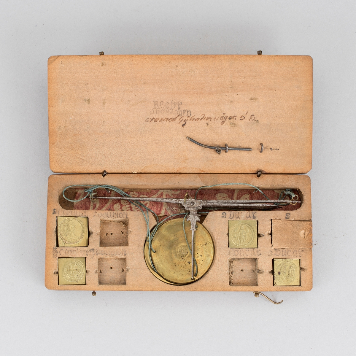 Myntvåg med etui. Vågbalk av järn med spatelformade ändar och åttaformade fästen. Visare av järn med fäste format som ett ankare. Kupiga vågskålar av mässing med inslaget mästarmärke, CM 8 i cirkel. I var skål tre hål för upphängning. Etui av valnötsträ med åtta fack för vikter var av ett med skjutlock.  Fyra kvadratiska stämplade vikter med myntbild för, Doublon, Carolin och Ducat samt mindre punsade balansvikter i skala 4 - 5. I övre delen av etuiet utskuret urtag för balansvåg. I etuiets lock en stålpinne för att lyfta vikterna ur sina fack. I lock stämpel med text Recht abgesogen (= riktigt avpassat) och annan otydd text på tyska. 10 delar totalt.