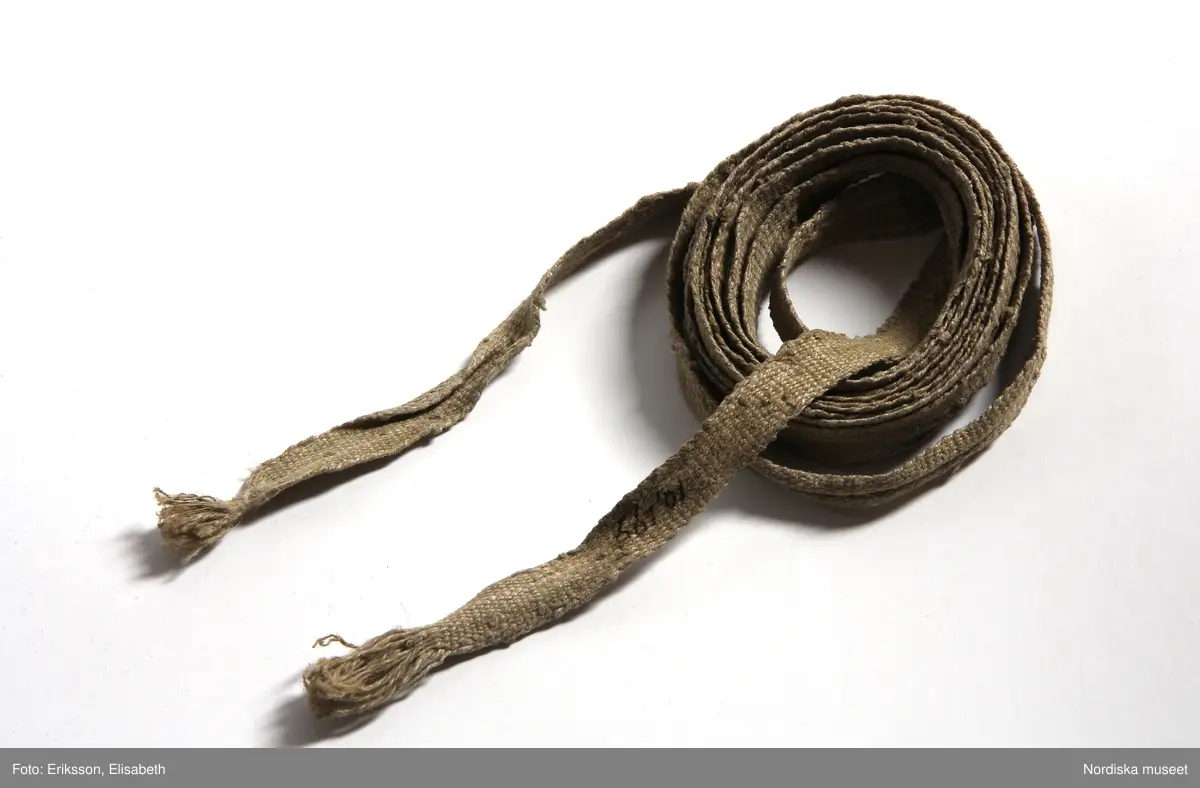 Kappa för kvinna av svart kläde, utan ärmar. Ålderdomlig typ.  Skuren i 6 våder, med nedpressade veck runtom.  Stripade rynkor upptill mot en  rundskuren besparing  av svart bomullssammet, som även fortsätter som kantgarnering längs framkanterna.   Svarta 2,5 cm breda  sidenband omger sammetsbesparingen och framkanterna. framkanterna är infodrade med  16cm bred remsa av grön  kypertvävd rask, ett glättat  tunt kamgarnsylle. I midjehöjd sitter i framkanternas insidor  ljusblå 28 cm långa bandöglor.  Kappan ofodrad förutom besparingen som är fodrad med oblekt linnelärft.  Knäpps högst upp i halsen med en stor hake och hyska av mässing. 
 Till kappan hör ett långt 2 cm brett  band litt b.  av grovt oblekt  hampa eller linblånor. har troligen använts vid förvaring av kappan och lindats runt för att behålla  vecken i fint skick. 
 Har tillhört  givarens mor Else Nilsdotter, men kan troligen vara ännu äldre från 1700-talet.
 Sigfrid Svensson skriver om sådana kappor använda som sorgdräkt då kvinnan bar  kappan över huvudet hopdragen så att bara ena ögat syntes. Går tillbaka på ett äldre sätt att bära kappor över huvudet även vid icke sorgtid på andra håll i Europa under 1500-och 1600-talen av kvinnor ur alla samhällsklasser. ( Skånes folkdräkter sid 273-274)