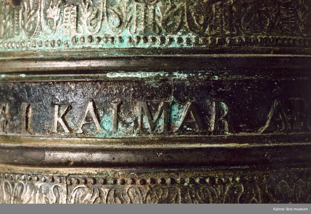 KLM 18217. Vällingklocka. Av malm. Dekor med gjutna lister samt text: GJUTEN AF I.P. FORSBERG I KALMAR ÅR 1857 samt ERIC SWARSS. Klockan är monterad på en metallstång, denna är målad i grå färg, som rostskydd, under senare tid.