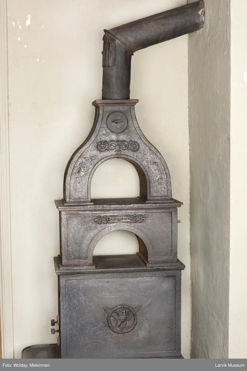 Jernovn i tre etasjer med muret fundament. Langs topp og rundbuet åpning rankeornament. Flatt askebrett med buede hjørner. 
1. etasje langside: Ørn i laurbærkrans 
1. etasje kortside: mrk.: FRITZØE