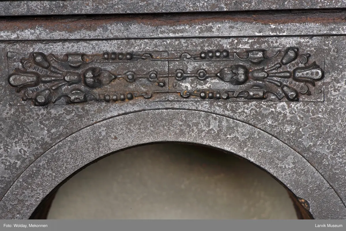 Jernovn i tre etasjer med muret fundament. Langs topp og rundbuet åpning rankeornament. Flatt askebrett med buede hjørner. 
1. etasje langside: Ørn i laurbærkrans 
1. etasje kortside: mrk.: FRITZØE