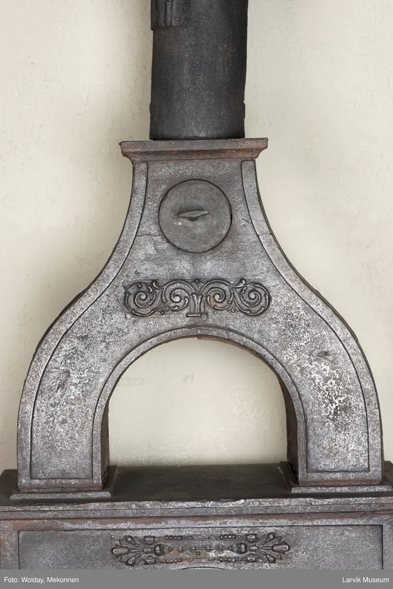 Jernovn i tre etasjer med muret fundament. Langs topp og rundbuet åpning rankeornament. Flatt askebrett med buede hjørner. 
1. etasje langside: Ørn i laurbærkrans 
1. etasje kortside: mrk.: FRITZØE