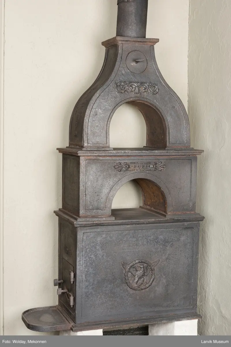 Jernovn i tre etasjer med muret fundament. Langs topp og rundbuet åpning rankeornament. Flatt askebrett med buede hjørner. 
1. etasje langside: Ørn i laurbærkrans 
1. etasje kortside: mrk.: FRITZØE