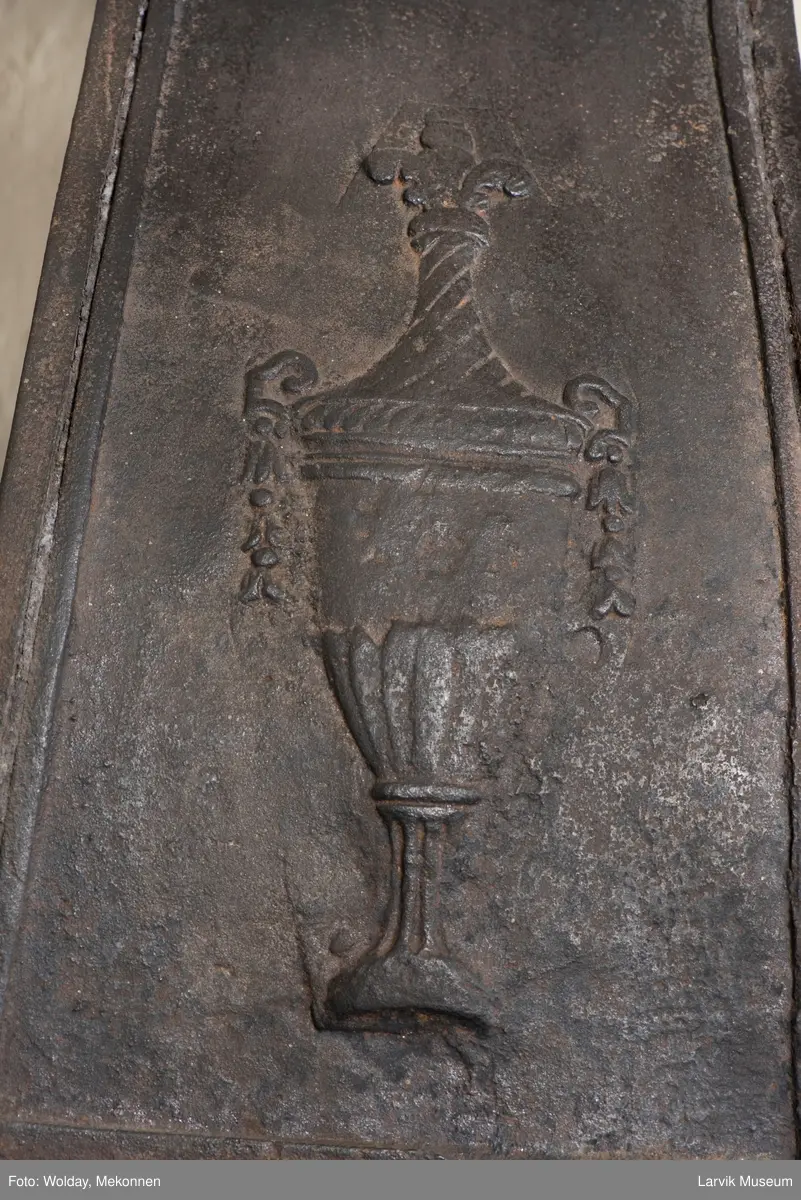 Jernovn i tre etasjer med muret fundament. Langs topp og rundbuet åpning rankeornament. Flatt askebrett med buede hjørner. 
1. etasje langside: Ørn i laurbærkrans 
1. etasje kortside: mrk.: FRITZØE