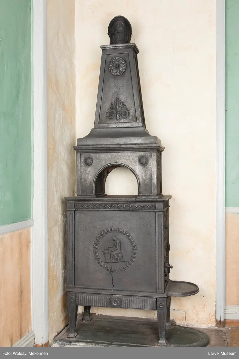 Tredelt , nederste etasje rektangulær fyrkasse/midtf.portalf/pyr.t. ben og sarg med kannelyrer og rosett i tre.  1.etasje langside: bladmedaljong, kvinne ved alter, øverst bølgemeander. 1.etasje kortside: bølgemeander, girlander og rosett 2.etasje langside: rundbuet åpning, i de øvre hjørner 2. etasje kortside: rosett i rektangel topp langside: palmett og rosett topp.Kortside: rosett i rektangel