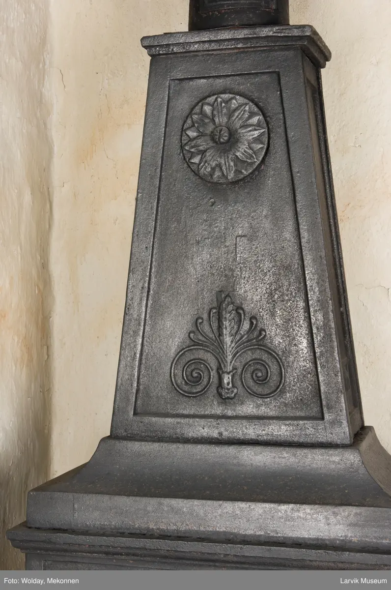 Tredelt , nederste etasje rektangulær fyrkasse/midtf.portalf/pyr.t. ben og sarg med kannelyrer og rosett i tre.  1.etasje langside: bladmedaljong, kvinne ved alter, øverst bølgemeander. 1.etasje kortside: bølgemeander, girlander og rosett 2.etasje langside: rundbuet åpning, i de øvre hjørner 2. etasje kortside: rosett i rektangel topp langside: palmett og rosett topp.Kortside: rosett i rektangel