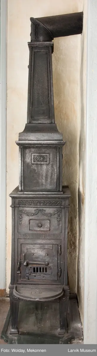 Tredelt , nederste etasje rektangulær fyrkasse/midtf.portalf/pyr.t. ben og sarg med kannelyrer og rosett i tre.  1.etasje langside: bladmedaljong, kvinne ved alter, øverst bølgemeander. 1.etasje kortside: bølgemeander, girlander og rosett 2.etasje langside: rundbuet åpning, i de øvre hjørner 2. etasje kortside: rosett i rektangel topp langside: palmett og rosett topp.Kortside: rosett i rektangel