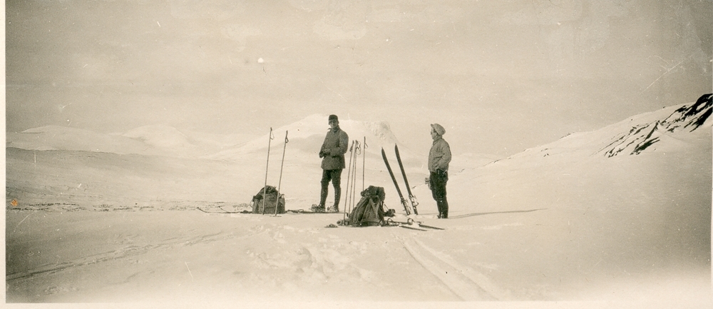 To menn står på snøen i skiløype. Høyfjell. Vinter. Skitur. Sekker og ski.