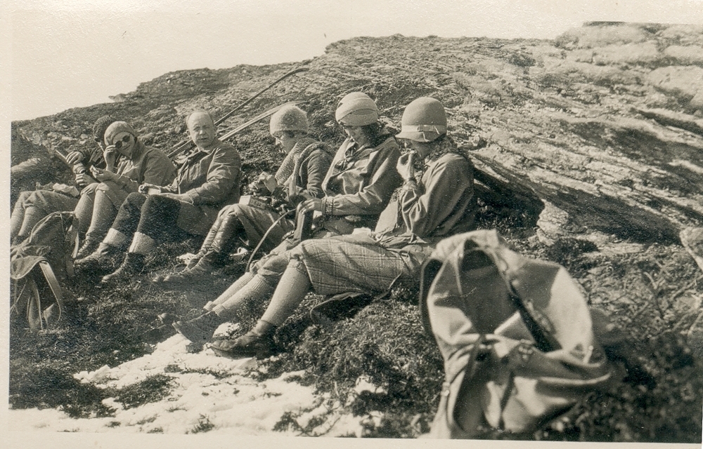 Matpause. Kafferast. Gruppe. Utendørs. Menn og kvinner sitter på bærmark, de har ski med seg. Sol. Fjell.