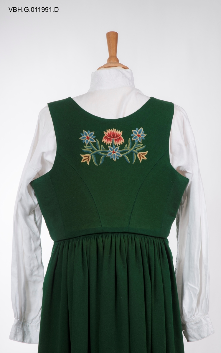 Broderier med blomstermotiver. Sjal og forkle med rutemønster.
Kjolen har blitt sydd ut i senere år med grønt bakstykke på 7 cm på hver side.