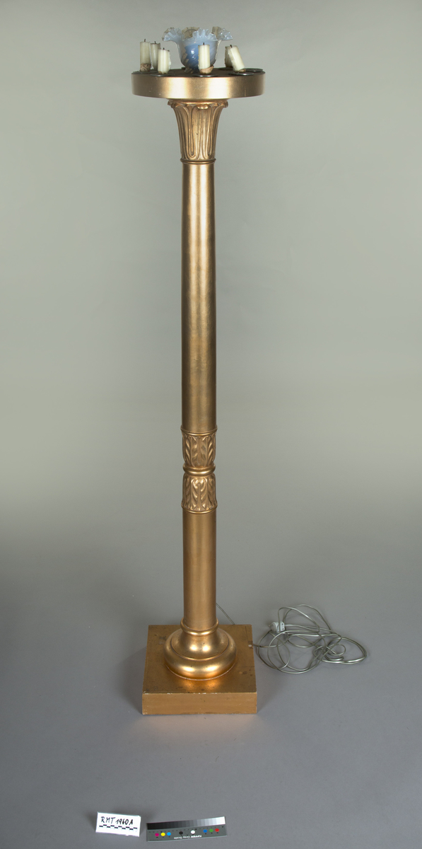 Forgylte gulvstående lamper for flere lys, både stearinlys og elektrisk. Hull til stearinlys langs kanten på hele topp-platen. En åpen lampeskjerm i glass omkranser det elektriske lyset midt på abakus, med bølgende kant (som en fakkel). Lampen er formet som en frittstående gresk søyle med base. Glatt utforming, med et område med bladutskjæringer like under midten, samt på kapitélet. Rund abakus og firkantet base.