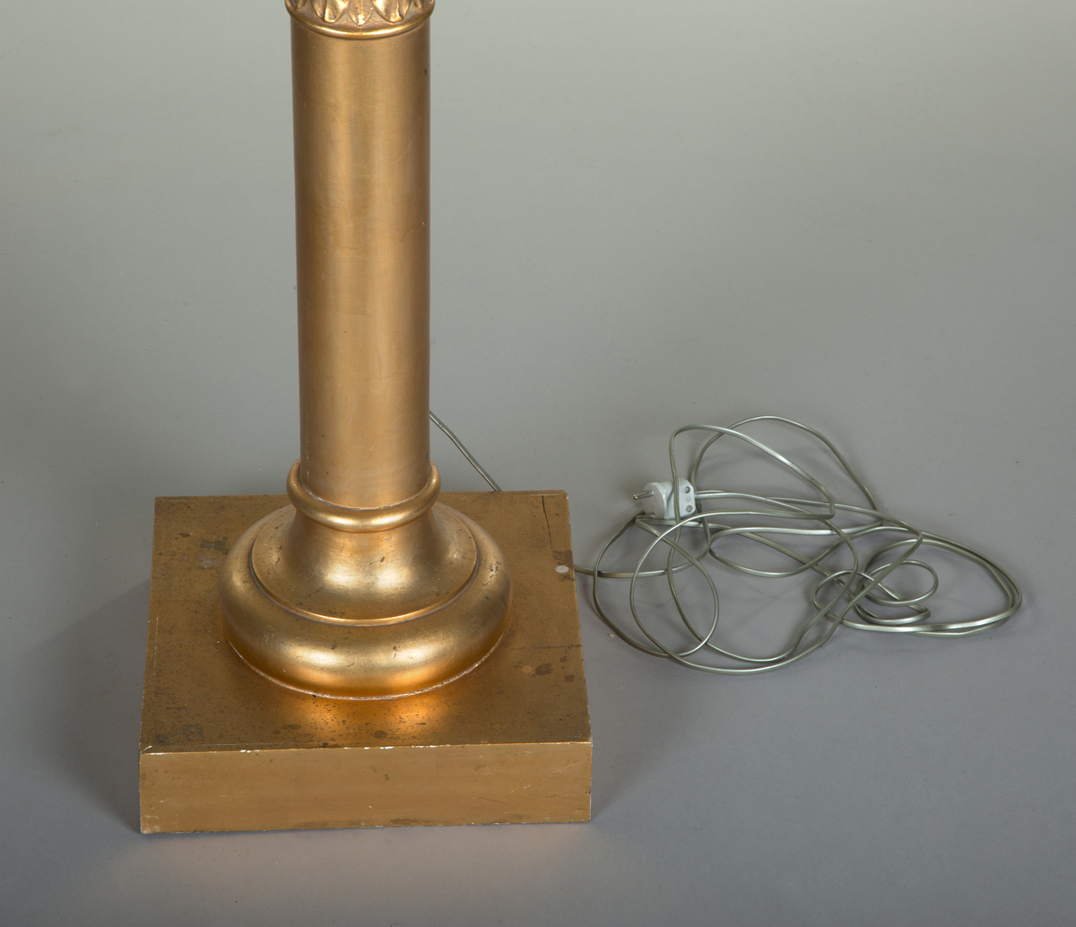 Forgylte gulvstående lamper for flere lys, både stearinlys og elektrisk. Hull til stearinlys langs kanten på hele topp-platen. En åpen lampeskjerm i glass omkranser det elektriske lyset midt på abakus, med bølgende kant (som en fakkel). Lampen er formet som en frittstående gresk søyle med base. Glatt utforming, med et område med bladutskjæringer like under midten, samt på kapitélet. Rund abakus og firkantet base.